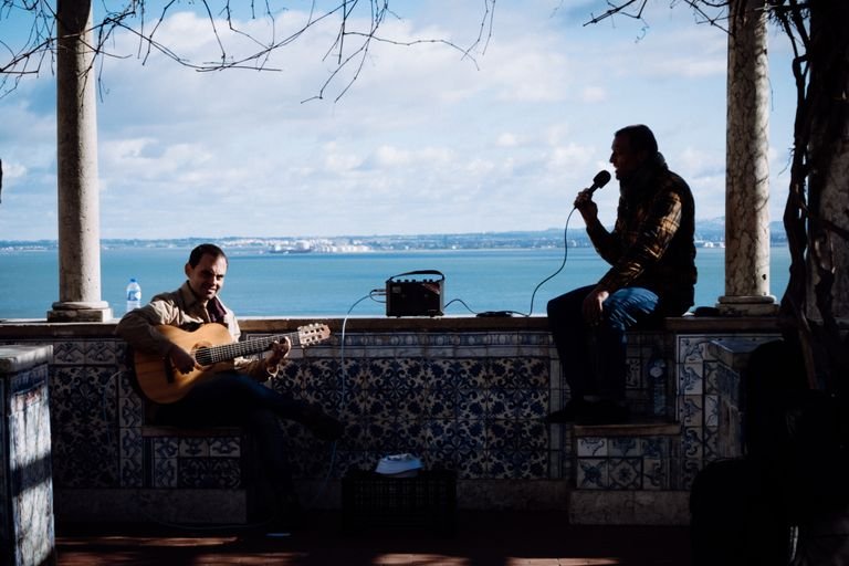 Two men perform music by the Lisbon seaside, one playing guitar and the other singing with a microphone.